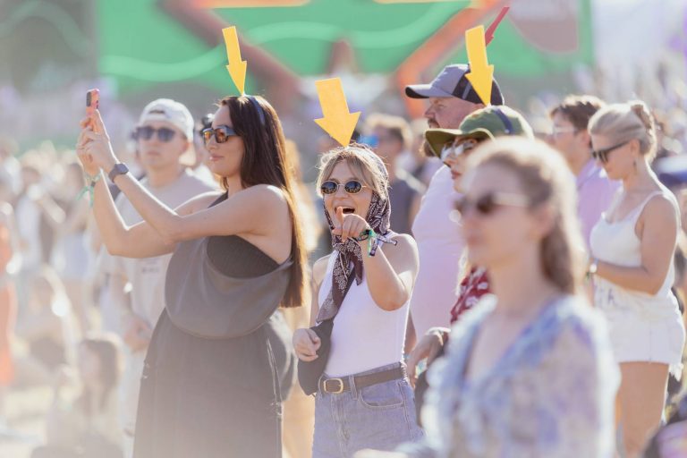 People enjoying an outdoor event with three individuals marked by arrows—a woman taking a selfie, a woman pointing and smiling, and a person in a green hat.