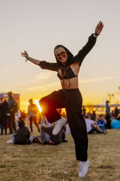 Smiling woman in black hoodie, black crop top, and sunglasses jumping with arms outstretched at outdoor event during sunset.