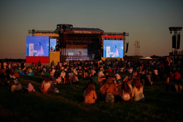 Large crowd sitting on grass watching a live music performance on stage at sunset during Opener Festival, with two large screens showing the singer at a piano.
