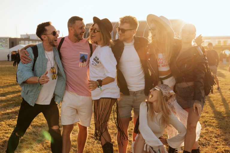 Group of six friends laughing and enjoying a sunny outdoor event, dressed in casual festival attire with sunglasses and hats.