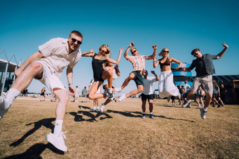 Group of young people jumping and having fun outdoors on a sunny day with a clear blue sky at a festival or event.
