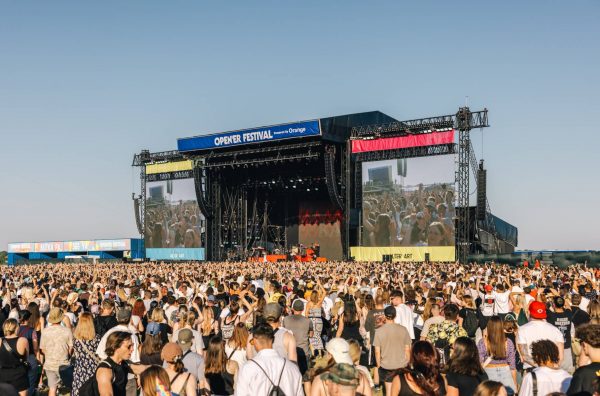 Large crowd of people at an outdoor music festival facing a stage with large screens displaying the audience, under a clear blue sky.