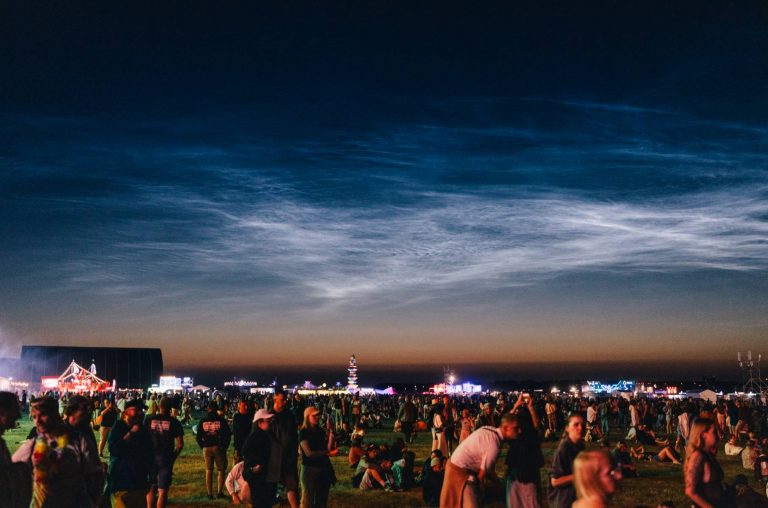 Crowd of people gathered at an outdoor festival during twilight with illuminated tents and rides in the background.