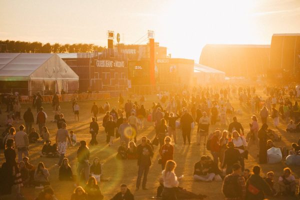 Crowd of people walking and sitting on grass at an outdoor event during golden hour with tents and structures in the background.