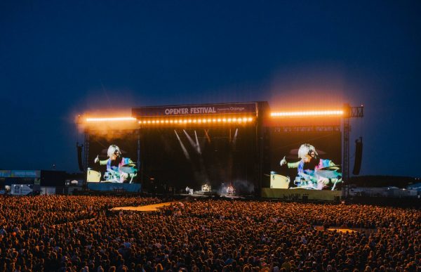 Large crowd watching a nighttime live performance on a brightly lit stage at the Open'er Festival with large screens showing a drummer.