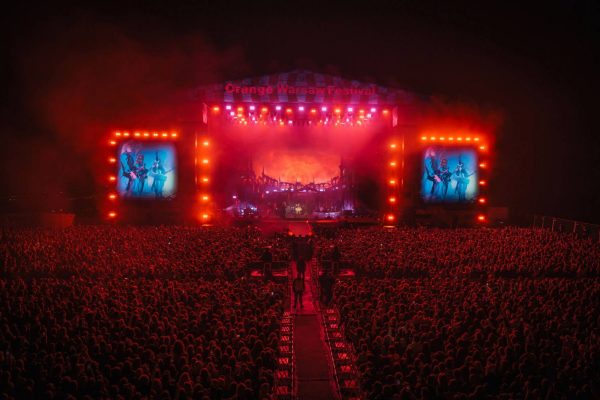 Large crowd at an outdoor night concert with bright red stage lights and two large screens displaying musicians.