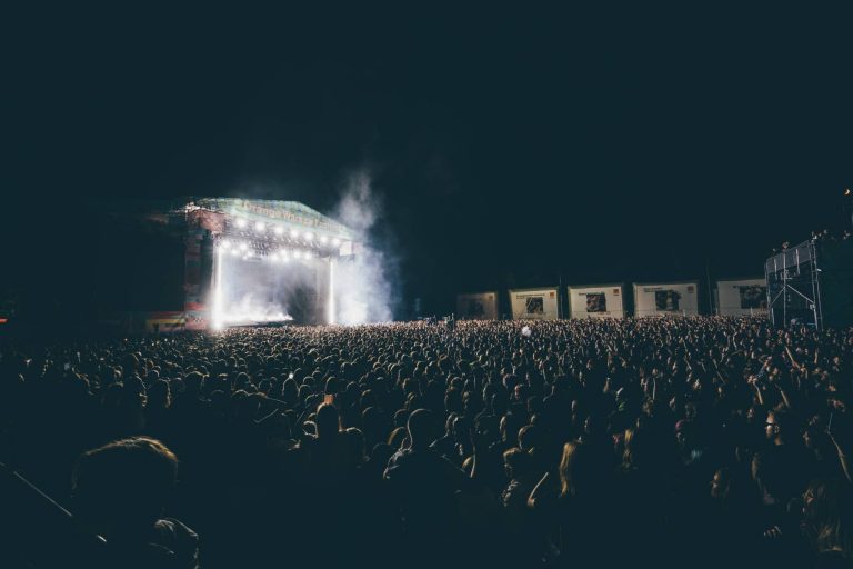 Large crowd gathered at night in front of a brightly lit outdoor concert stage with smoke effects and banners in the background.