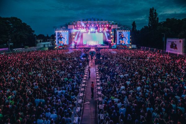 Large crowd gathered at night facing a brightly lit stage with screens displaying a performer at the Orange Warsaw Festival.