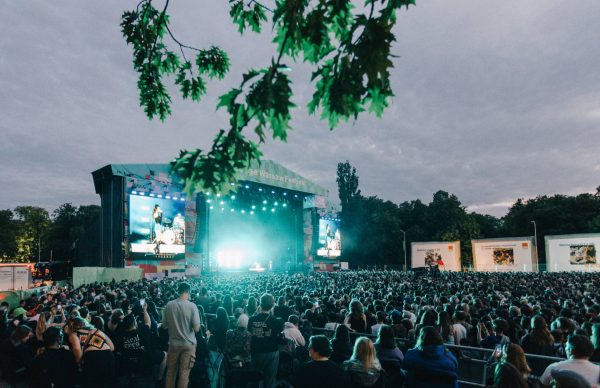 Large crowd of people watching a brightly lit concert stage at an outdoor festival under a cloudy sky with tree branches hanging overhead.