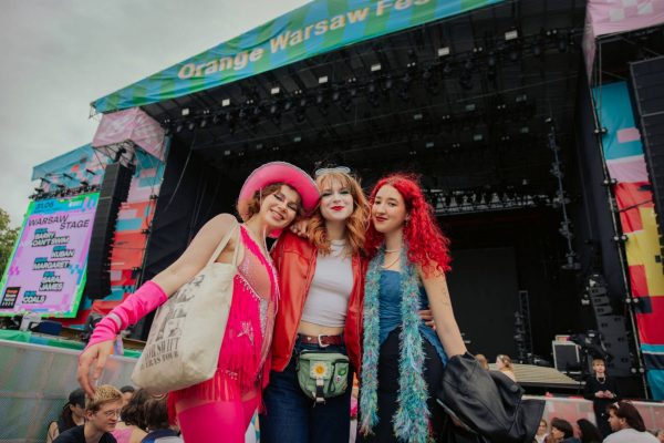 Three young women in vibrant festival outfits posing and smiling in front of a large outdoor concert stage with a colorful banner reading 