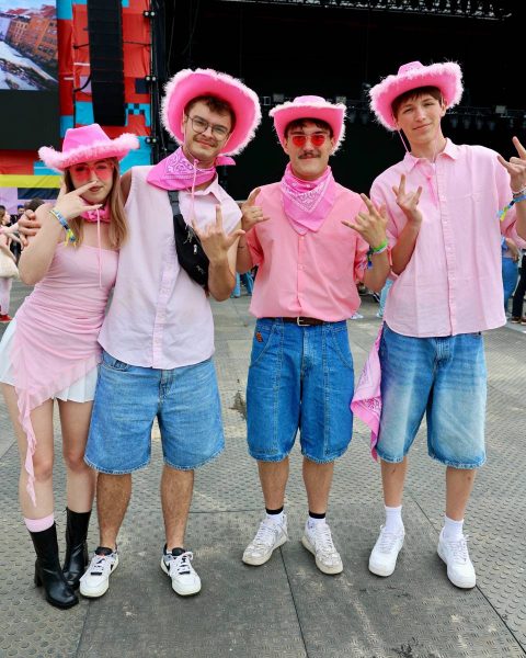 Four young people wearing pink hats, pink tops, and denim shorts posing with hand signs at an outdoor event.