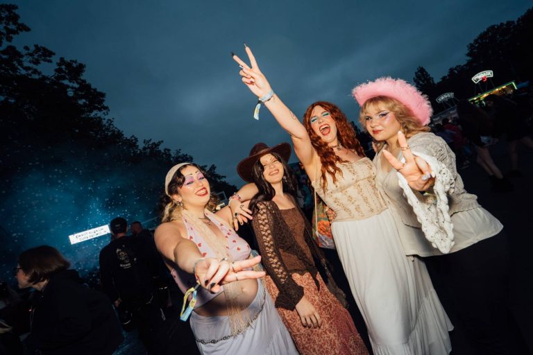 Four women in festival attire smiling and posing with peace signs at an outdoor evening event.