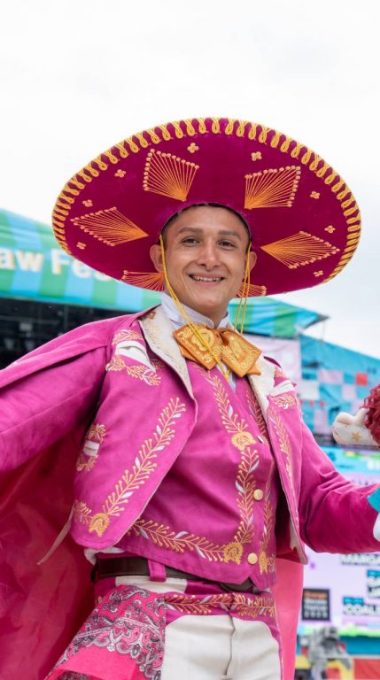 Man smiling wearing a bright pink traditional charro suit and large embroidered sombrero at an outdoor event.