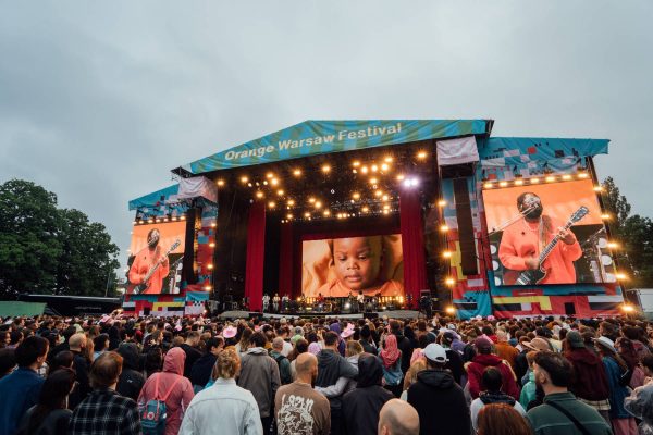 Large crowd watching a live music performance on a brightly lit stage at the Orange Warsaw Festival with screens showing a musician and a baby.