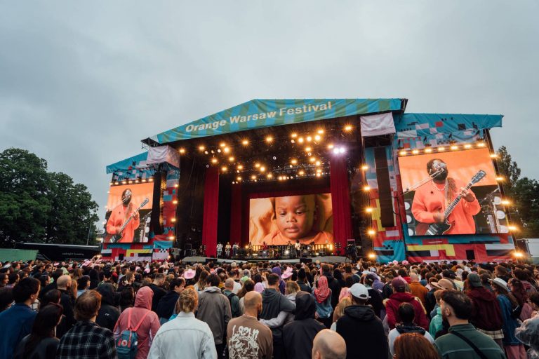 Large crowd watching a live music performance on a brightly lit stage at the Orange Warsaw Festival with screens showing a musician and a baby.