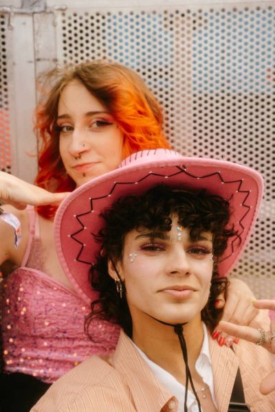 Two young people with party makeup, one with curly hair wearing a pink cowboy hat and striped beige shirt, the other with red-orange hair in a pink sequined top