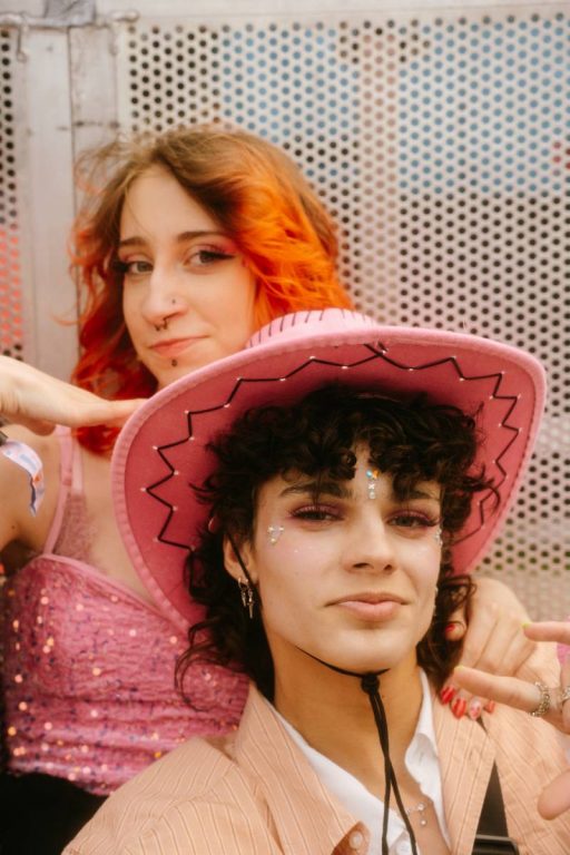 Two young people with party makeup, one with curly hair wearing a pink cowboy hat and striped beige shirt, the other with red-orange hair in a pink sequined top