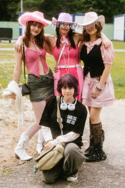 Group of four young people posing outdoors, three standing wearing pink outfits and cowboy hats, one kneeling holding a bag and wearing headphones around their neck.