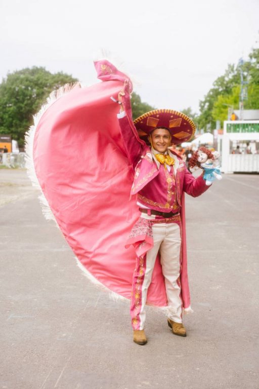 Man wearing a vibrant pink and white traditional charro suit with a large sombrero, holding a puppet and raising a flowing cape outdoors.