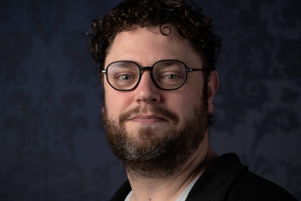 Man with curly hair, glasses, and a beard smiling slightly against a dark patterned background.