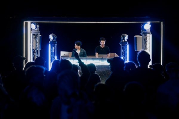Two DJs performing on a lit stage with laptops and spotlights, facing a crowd at night.