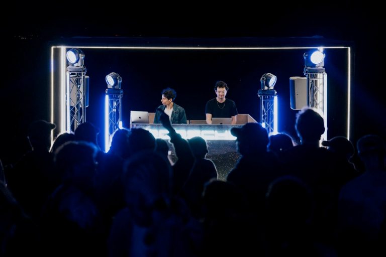 Two DJs performing on a lit stage with laptops and spotlights, facing a crowd at night.