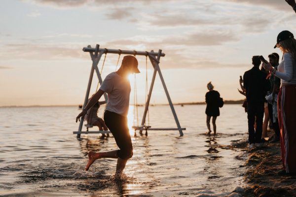 Person splashing water with one leg in shallow beach water near a wooden swing set at sunset with others standing on the shore nearby.