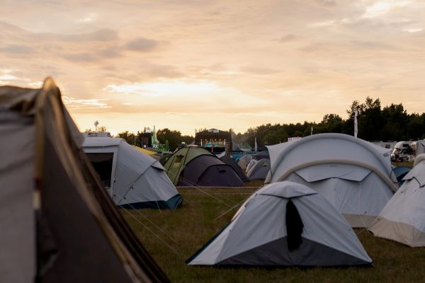 Various tents set up on grassy field at sunset with a music festival stage in the background and trees beyond