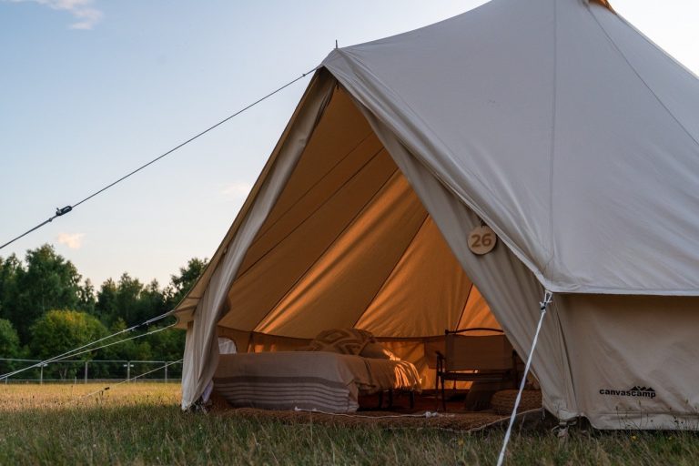 Large canvas tent with a bed and chair inside, set up in a grassy field near trees during daylight.