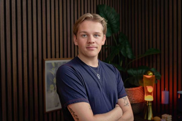 Young man with light hair and a mustache wearing a navy blue t-shirt and a silver female symbol necklace, standing with arms crossed indoors.