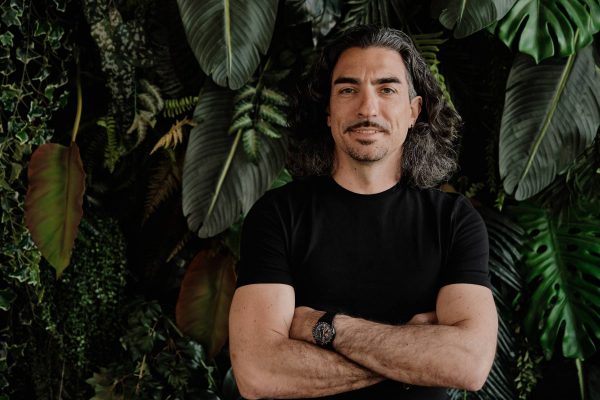 Man with long wavy hair and mustache wearing a black t-shirt and watch, standing with arms crossed in front of large green leaves.