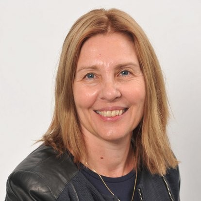Smiling woman with shoulder-length blonde hair wearing a black leather jacket over a dark top against a plain background.