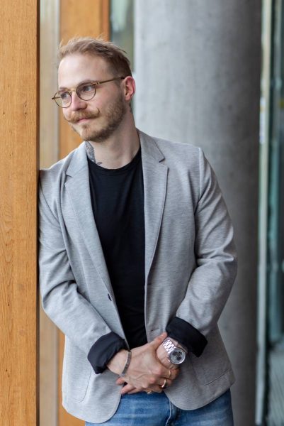 Man with glasses, mustache, and beard wearing a gray blazer, black shirt, and jeans, standing by a wooden pillar indoors.