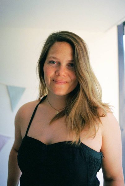 Smiling woman with light brown hair wearing a black halter top and a small round pendant necklace indoors.