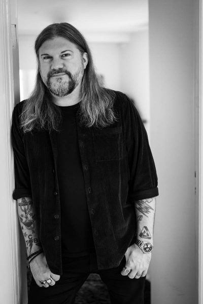 Man with long hair and beard wearing a black shirt, showing tattooed arms, rings, and a watch, standing by a doorway in black and white.