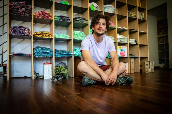 Man with curly hair wearing a white t-shirt and shorts sitting cross-legged on a wooden floor in front of shelves stocked with folded colorful t-shirts.