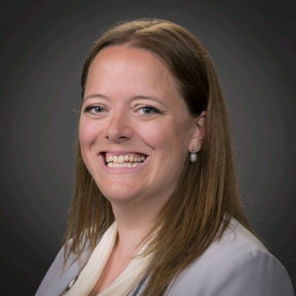 Smiling woman with long brown hair wearing a light gray blazer and white blouse against a dark background.