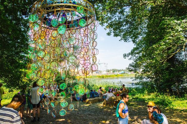 Colorful hanging circular glass decorations with people sitting on hay bales and socializing outdoors near a river under trees.