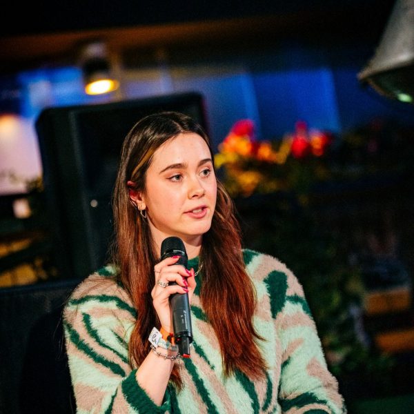 Young woman with long brown hair, wearing a green and beige striped sweater, speaking into a microphone indoors.