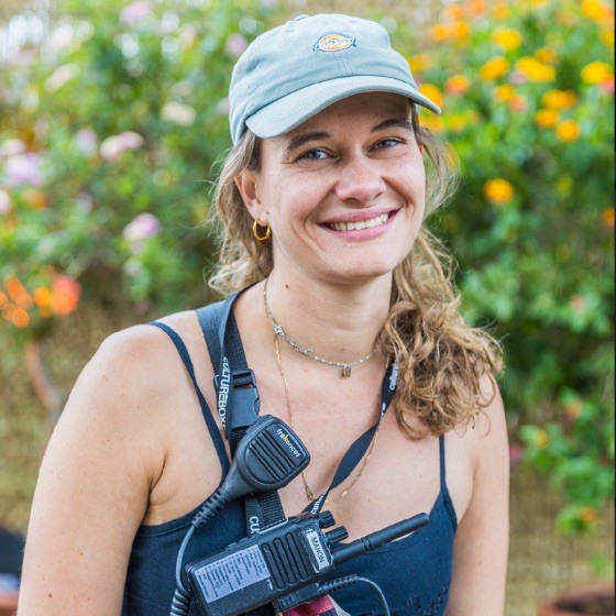 Smiling woman wearing a blue cap and tank top with a walkie-talkie clipped to her strap outdoors with blurred flowers in the background