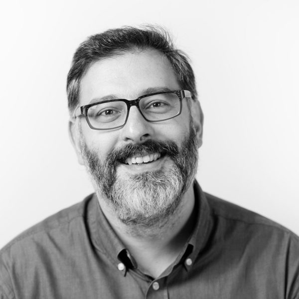 Bearded man wearing glasses and a button-up shirt smiling against a plain white background in black and white.