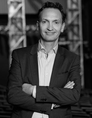 Man in a suit with arms crossed, smiling in a black and white indoor setting.