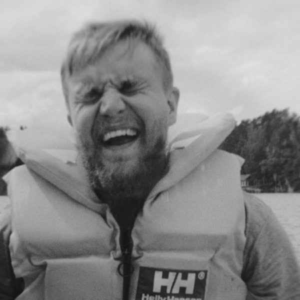 Man wearing a life jacket smiling with eyes closed near a body of water with trees in the background