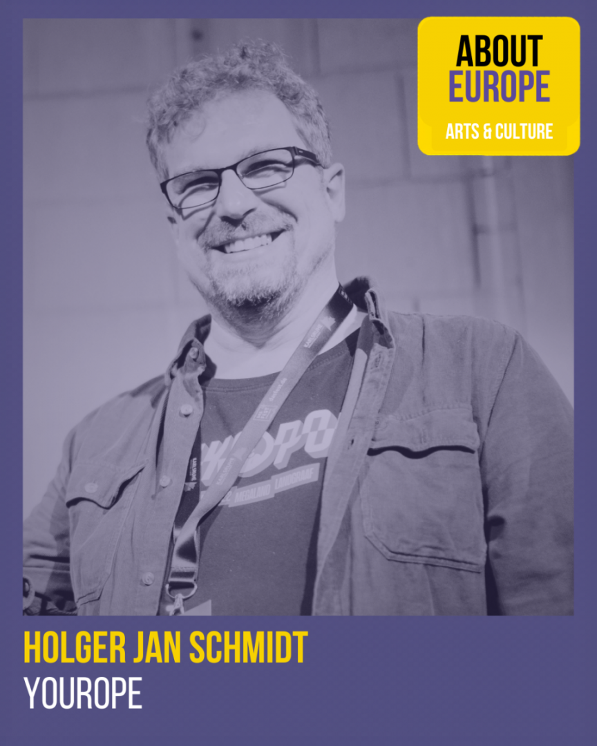 Man with glasses and curly hair smiling, wearing a shirt and jacket with a lanyard around his neck, labeled Holger Jan Schmidt, Yourope.