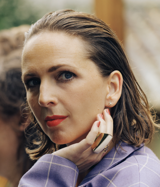 Woman with short wet hair wearing a purple checkered blazer, red lipstick, a gold ring, and a lightning bolt earring, resting her hand on her chin.