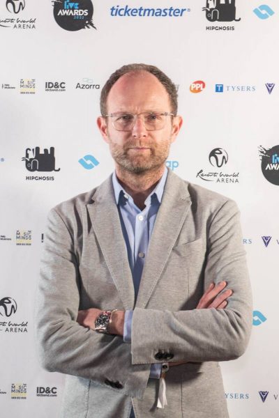 Man with glasses and beard in light gray blazer standing with arms crossed in front of a white backdrop with logos including ticketmaster and Resorts World Arena