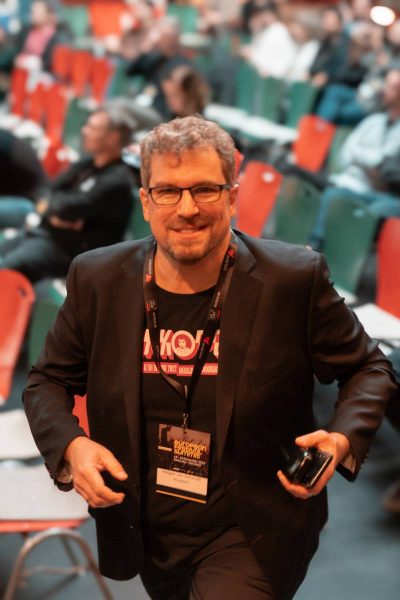Man wearing glasses, black blazer, and conference badge walking in an auditorium with colorful empty seats in the background.