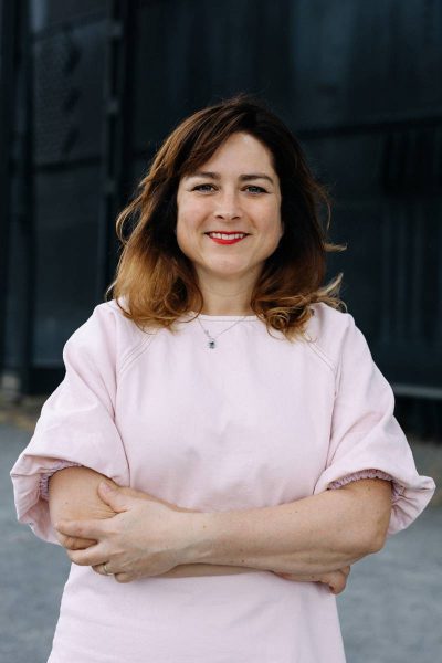 Smiling woman with ombre hair wearing a light pink blouse and red lipstick, standing outdoors with arms crossed.