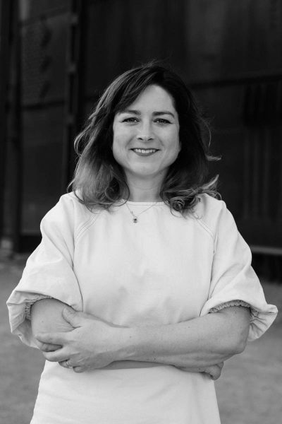 Smiling woman with medium-length wavy hair wearing a light-colored shirt with rolled-up sleeves and arms crossed.